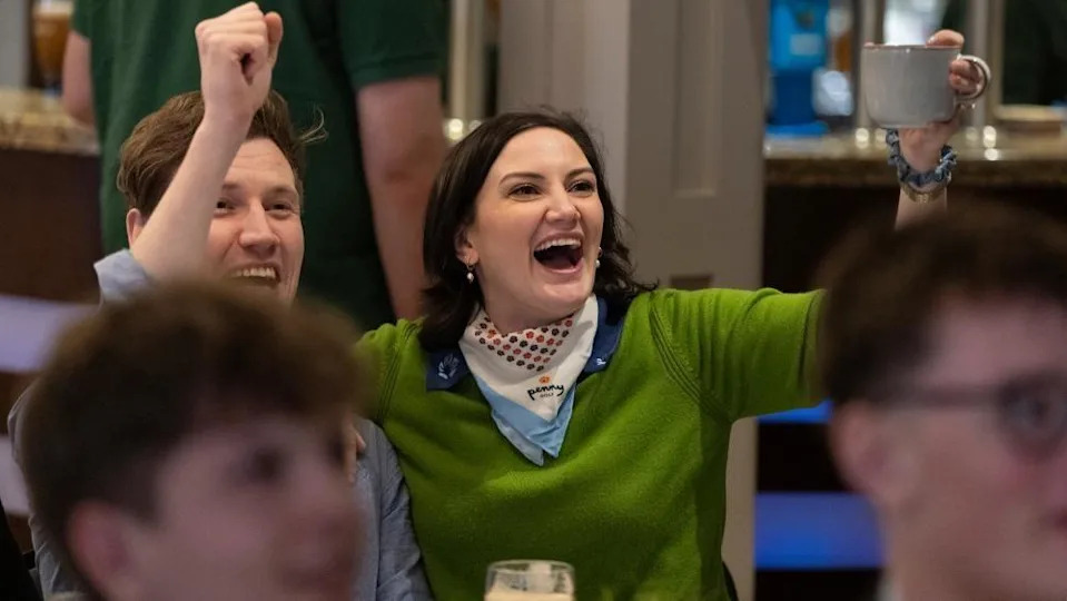 Leah James  is wearing a green jumper and cheering with hands raised aloft in a golf club supportive of Rory McIlroy. She is sitting beside a man who is also cheering. She has a silk scarf around her neck and is holding a mug in her left hand.