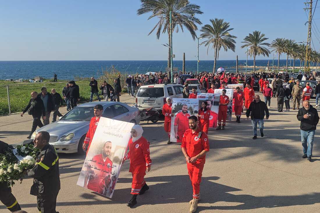 Mourners hold a portrait of Youssef Assaf, a Lebanese Red Cross volunteer killed during a rescue mission in southern Lebanon, at his funeral. Many people are wearing Red Cross uniforms as they walk along the coast of the Mediterranean Sea.